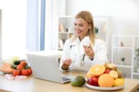 Dietitian holding supplement bottles during virtual St Petersburg nutritionist services consultation with fresh fruits and vegetables on desk.